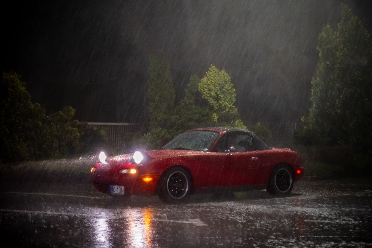 The Art of Drawing Readers In: Your attractive post title goes here Red Mazda Miata parked in heavy rain at night, showcasing vehicle lights and wet roads.